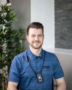 Adult white male with short brown hair is sitting with a blue short-sleeved polo shirt and a ListenIR receiver and neck loop around his neck.