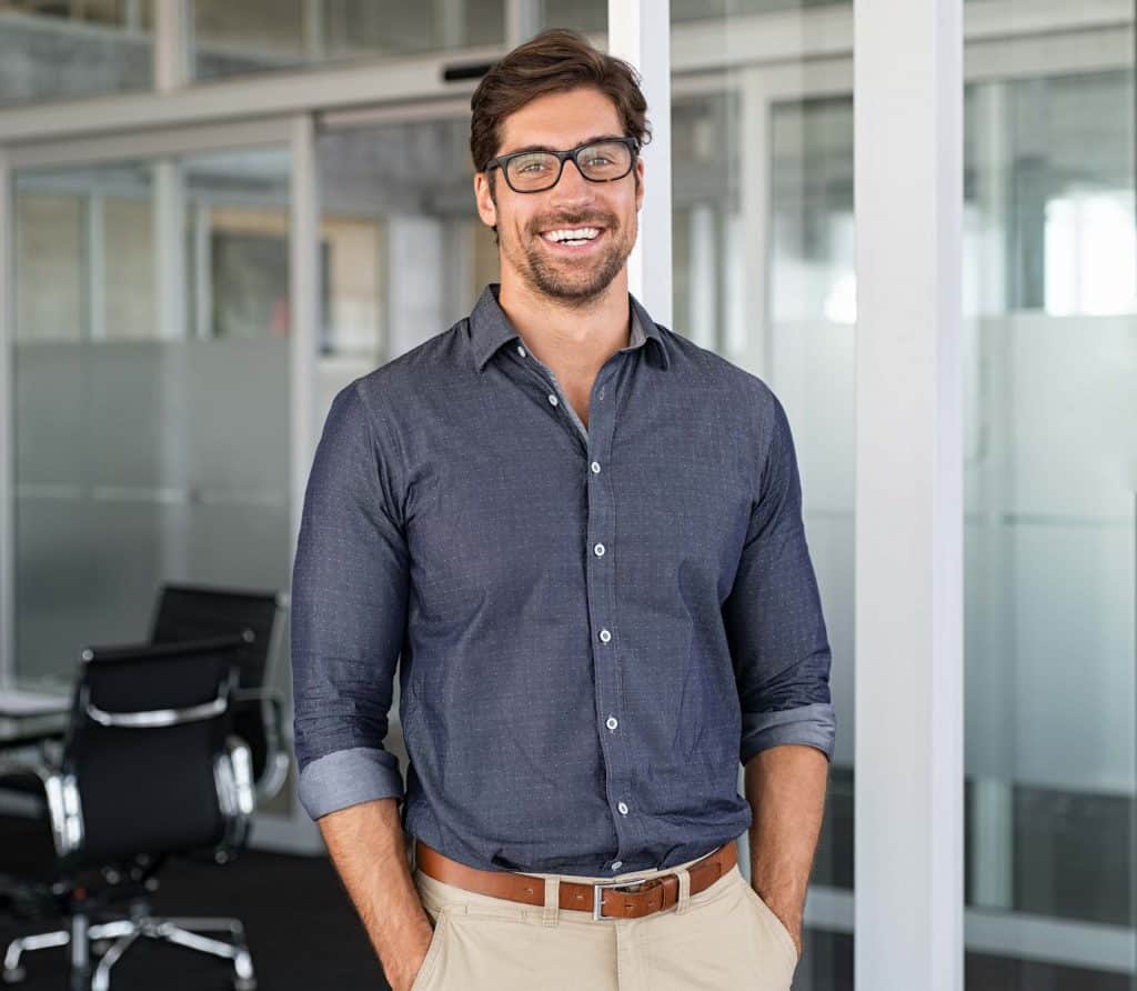 Bell'aspetto, sorridente e sicuro di sé, un uomo di 30 anni in un ambiente d'ufficio, con indosso occhiali con montatura nera, capelli castano scuro corti con la riga di lato e piumati, con peli facciali corti, denti molto bianchi, indossa una camicia con colletto blu scuro con un fantasia chiara, cintura marrone e pantaloni color cachi con le mani nelle tasche dei pantaloni.