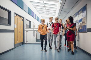 Un gruppo di sei giovani uomini adulti che camminano lungo una sala del campus con una donna afroamericana adulta vestita in modo professionale, tutti indossano dispositivi ListenTALK al collo con auricolari.