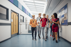 Grupo de seis hombres adultos jóvenes caminando por un pasillo del campus con una mujer afroamericana adulta vestida profesionalmente, todos con dispositivos ListenTALK alrededor del cuello con auriculares.