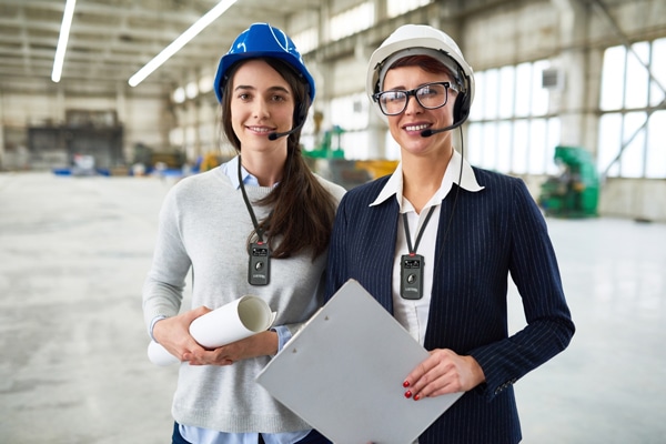 Two females wearing blouses, hard hats, holding a poster and clipboard, and wearing Listen products around their neck.