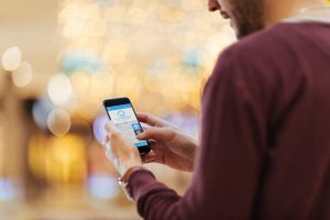 bottom half of a guy's face and the top portion of his torso, wearing a maroon, long-sleeved sweater, a brown wristband watch, and looking at his phone which is displaying the Listen EVERYWHERE app.