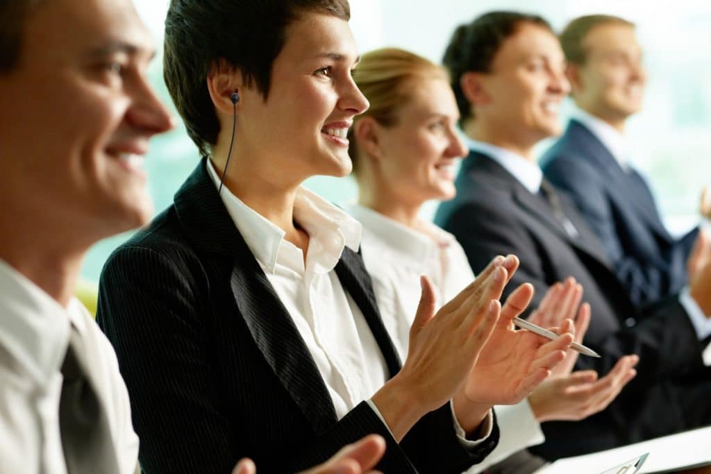 Row of professionally dressed white people clapping and smiling with the focus of the photo on young, brunette, short-haired woman wearing ear buds