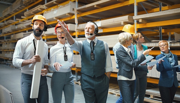 Closeup front view of mixed age people of industrial design department at a factory. They are walking in an aisle between pallet racks stacked with chipboard, MDF and plywood. There are three men and four women. They are using ListenTALK to communicate.