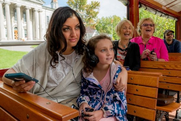 Jeune femme aux cheveux noirs et sa fille d'environ 8 ou 9 ans écoutant de l'audio avec différents casques, assises sur un chariot de tournée avec des bancs en bois, des côtés en plein air et d'autres touristes, deux femmes plus âgées, riant en arrière-plan.