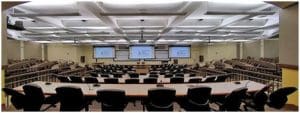 Grande salle de classe, bien éclairée, avec tables et chaises en rangées, trois grands moniteurs vidéo à l'avant de la salle.