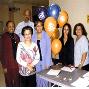 Six adults standing at a table with papers on it and orange, blue, and purple balloons.