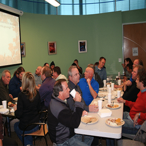 Listen Technologies-Mitarbeiter sitzen und genießen ihr Mittagessen an langen Tischen in einem grün gestrichenen Raum.