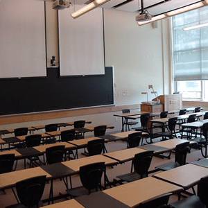 Empty classroom showing desks, chairs, a bright window with the shade partially down, fluorescent lighting, AV cameras and projectors installed on the ceiling and the wall, a long chalkboard, and a teaching pulpit.