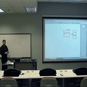 Classroom view from a students perspective with a teacher/presenter at the front of the classroom with a screen projector displayed