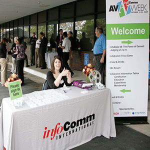 Mesa de bienvenida con un mantel blanco con InfoComm International impreso en el frente. El banner desplegable ubicado a la derecha dice AV Week, Welcome, con un texto ilegible más pequeño. Amable mujer sonriente sentada a la mesa con multitudes errantes de personas en el fondo.