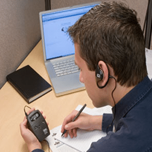 young man with short, brown hair working at a desk writing on paper looking at an assistive listening receiver with a universal ear speaker over one ear