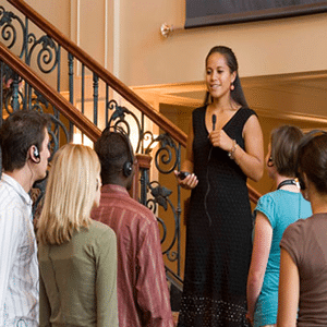 A woman in a black sleeveless dress standing on a staircase with elaborate iron work is speaking into a microphone to a group of people using headphones