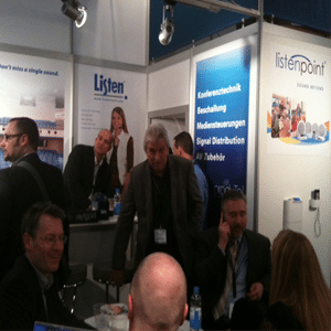 Salle d'exposition animée avec des groupes de personnes debout et d'autres groupes assis sur un stand de Listen Technologies