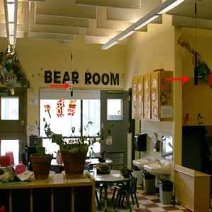 Early learning classroom with big bright window, small table with small chairs, two sinks for a handwashing station with cupboards above, and a long counter with learning accessories with red and white tile floors in a checker pattern