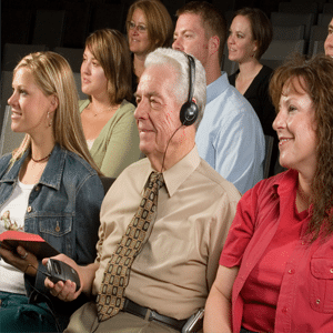 Group of people sitting as an audience with an older gentleman in a dress shirt and a tie, wearing a headset and holding an assistive listening receiver in his hand