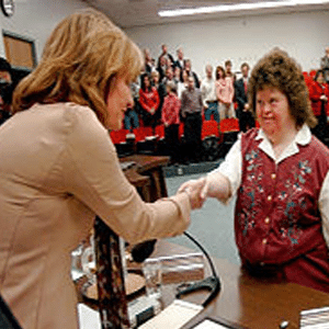 Brown-haired woman in suit jacket is shaking the hand of a young woman in a collared dress shirt and button-down sweater vest