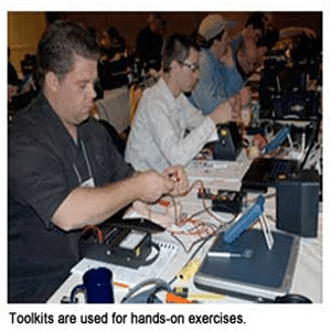 A long table with electronic equipment and men sitting in front of them doing something with their hands with the wiring