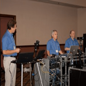 Photo de trois messieurs blancs plus âgés en chemises bleues et kakis devant un équipement de sonorisation