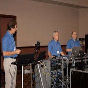 Photo of three white, older gentlemen in blue shirts and khakis in front of sound equipment