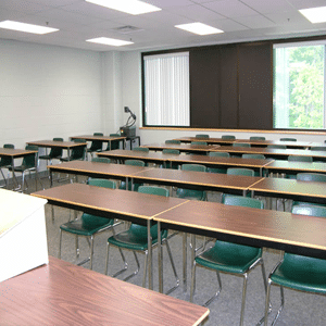 Room with long rectangular tables and green classroom-style chairs behind them with a podium at the front of the room.