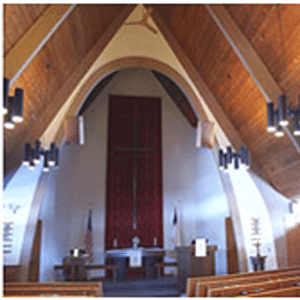 The view of a church sanctuary with a large cross on the wall behind a pulpit with two flags on either side, empty pews, and vaulted ceilings