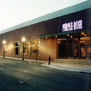 Picture of a non-descript brown brick building at dusk with lights on and the name Purple Rose in purple lights over the entrance