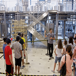Tour of a brewery - a group of about 20, casually dressed, people wearing headphones, facing one guy dressed in black jeans and gray t-shirt, also wearing a headset with a microphone and pointing to something behind him.