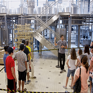 Besichtigung einer Brauerei - eine Gruppe von etwa 20 Personen, leger gekleidet, Leute mit Kopfhörern, einem Mann in schwarzer Jeans und grauem T-Shirt gegenüber, der ebenfalls ein Headset mit Mikrofon trägt und auf etwas hinter ihm zeigt.