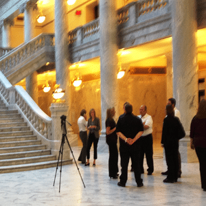 All'interno dello Utah State Capitol Building con pavimento in marmo, scala in marmo e grandi colonne di marmo con un gruppo di persone in piedi intorno a quella che sembra essere una telecamera montata su un treppiede