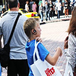 En la calle con dos padres que acompañan a un preadolescente rubio sucio que lleva un parlante, una camiseta azul y una bolsa blanca con el logo de Seattle Bites.