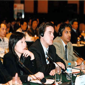 a group of people in a room paying attention with the focus on three people seated at a long table with microphones in front of them, two are wearing headsets.