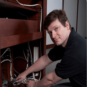 Brown-haired, blue-eyed 30-something man wearing a short-sleeved, black polo, squatting with both hands on wires in a nicely finished wooden shelving unit, looking at the camera.