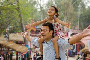 Father with daughter on his shoulders at venue. Both arms are outstretched