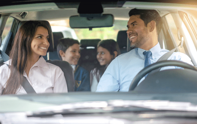 Family in a car driving. On the right is the dad with dark hair, wearing a light blue shirt and dark blue tie. On the left is the mother with dark hair, wearing a light pink blouse. In the backseat of the car are the daughter (right) and son (left) looking at each other, smiling. 