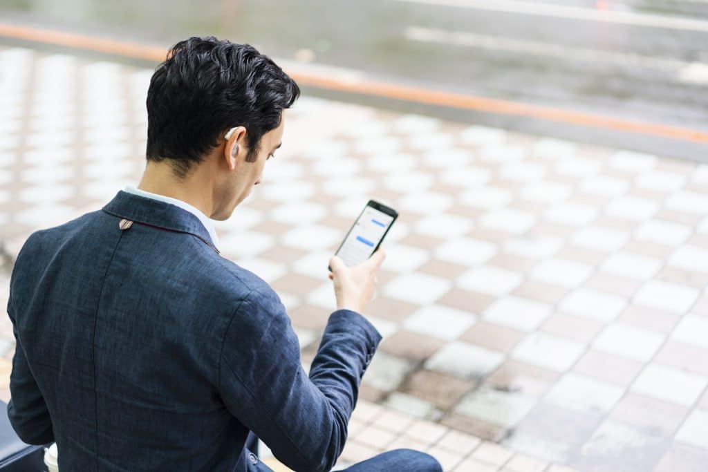 Jeune homme d'affaires métis avec des aides auditives assis sur un banc et tapant un message sur son téléphone en attendant un bus