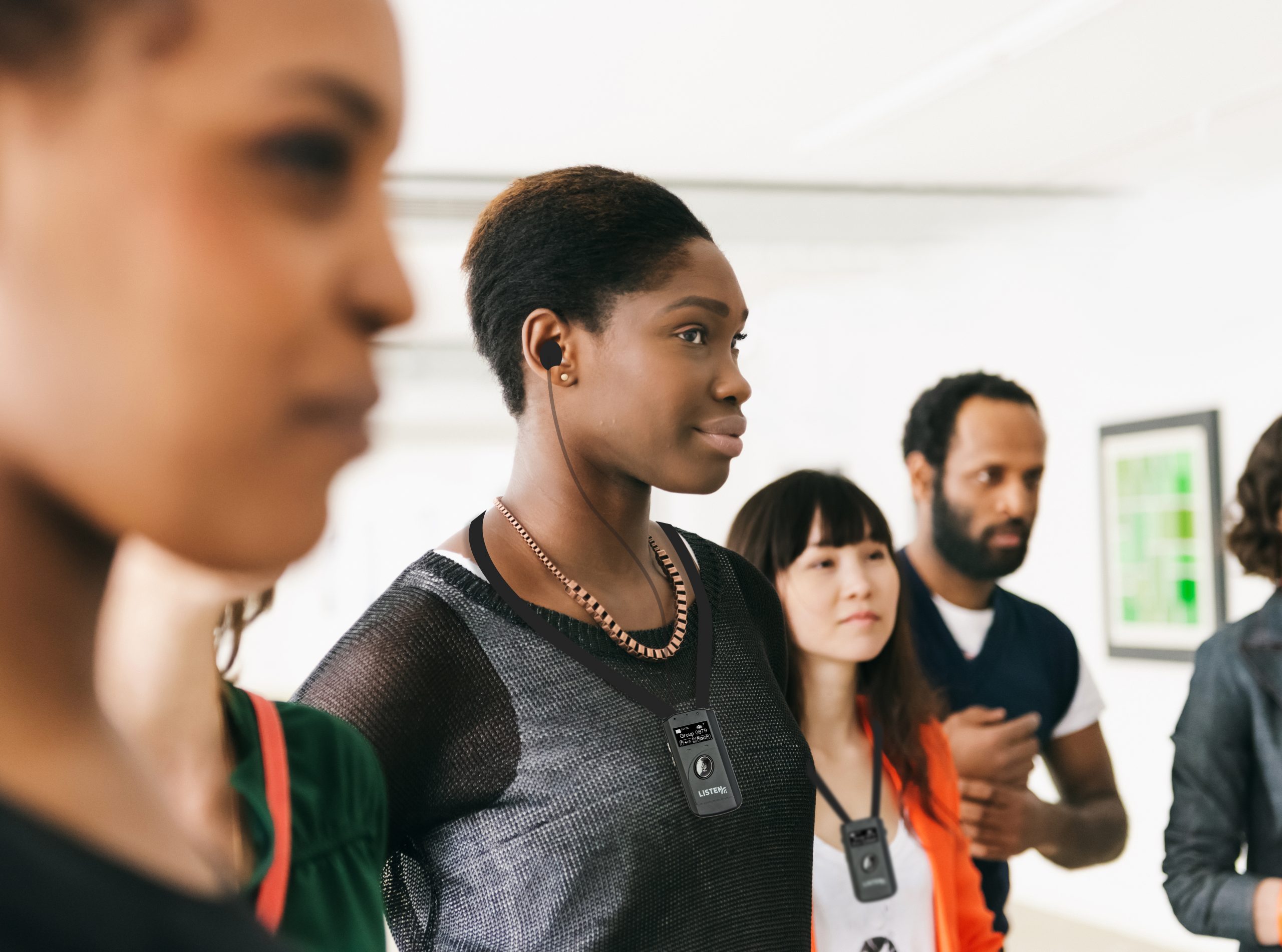 Mixed group of visitors standing and listening during a gallery opening in Berlin, Germany. Focus is on a young black african woman smiling and wearing earbuds. Horizontal shot of people with artwork in the background hanging on the wall.