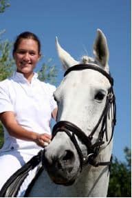 Cloe up of a young woman with long brown hair that's pulled back, wearing a white short sleeved button-down top, riding a white horse with a black bridle.