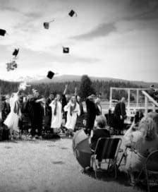 Foto en escala de grises de un grupo de graduados de secundaria de pie afuera y lanzando sus gorras al aire en celebración frente a un grupo de personas en sillas