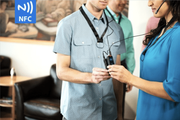 A man in a light blue short-sleeved button down shirt tapping his ListenTALK devices to pair it with a brunette woman in a blue blouse. There is also a logo in the top left-hand corner that says NFC and has waves on it. NFC stands for near-field communication.