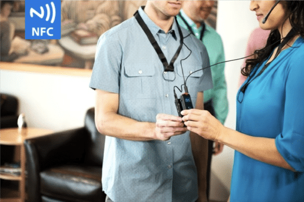 Un hombre con una camisa azul claro de manga corta con botones tocando sus dispositivos ListenTALK para emparejarlo con una mujer morena con una blusa azul. También hay un logotipo en la esquina superior izquierda que dice NFC y tiene ondas. NFC significa comunicación de campo cercano.