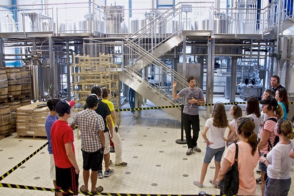 Tour of a brewery - a group of about 20, casually dressed, people wearing headphones, facing one guy dressed in black jeans and gray t-shirt, also wearing a headset with a microphone and pointing to something behind him.