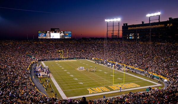Outdoor football field and arena, Lambeau Field, at dusk, with players on the field and on the sidelines, fans in the arena, the lights are on and there is a large digital display at the end