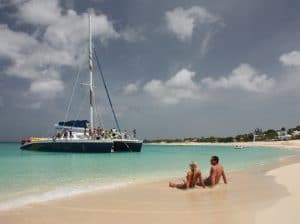 Pareja de mediana edad sentada en una playa tropical admirando un velero que llega a la orilla.