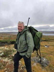 Halvard Eriksen sonriendo, de pie al aire libre en un hermoso paisaje con nubes y hierba cubierta de musgo, con un abrigo verde, una mochila pesada y su rifle de caza.