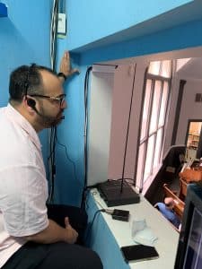 Big man with glasses and dark hair sits in a brightly painted blue room above and at the back of the church sanctuary with Listen Technologies' ListenRF system for interpretation