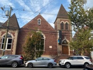 An older church building with red bricks and brown roofing. Three cars are parked out in front on the street. 