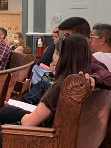 A young adult woman sits in a pew with a headset on listening to the services in English