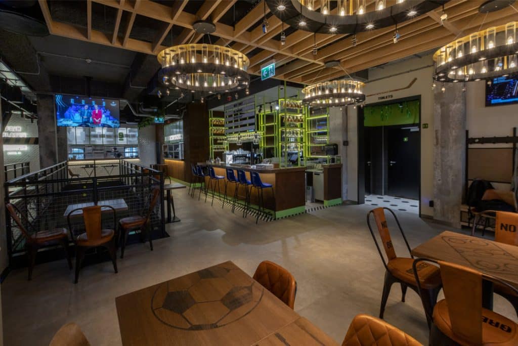 Restaurant and bar area with chandelier lighting, table and chairs seating, and a bar with blue high stools. Guests can tune into the Wi-Fi Audio Streaming at this sports bar.