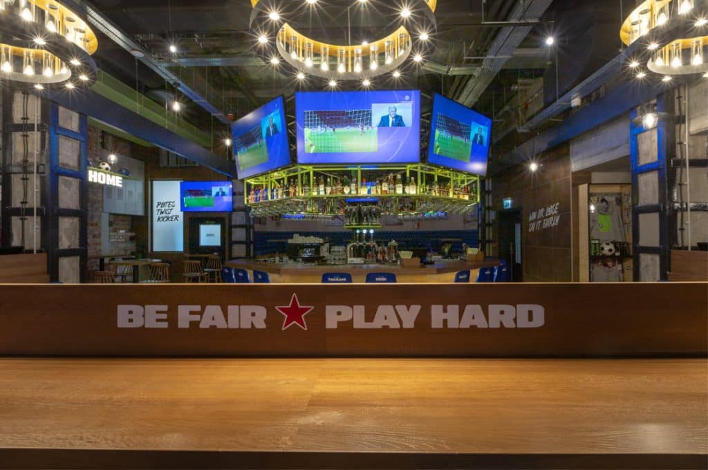 Grand espace bar avec les mots Be Fair Play Hard sur un panneau. De grands écrans de télévision sont suspendus au-dessus du bar avec des matchs de football sur les écrans.